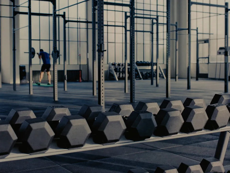 Rows of dumbbells on a rack in a bright training facility, with an athlete setting up a barbell behind.