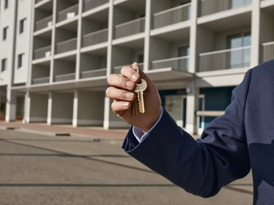 A property manager holds a set of keys in front of a well-kept apartment building.